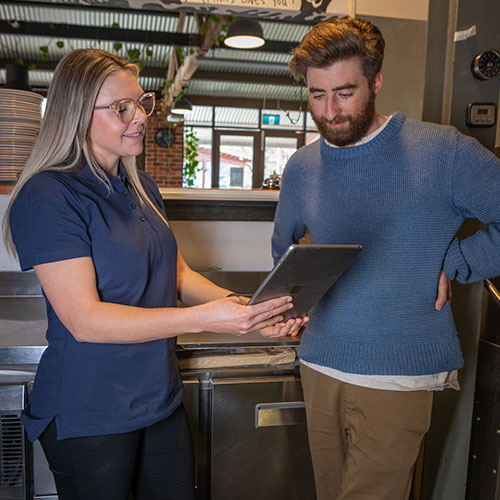 A business energy assessor in a blue top pointing to an iPad. A man is looking at the iPad and smiling. They're both standing in a commercial kitchen.